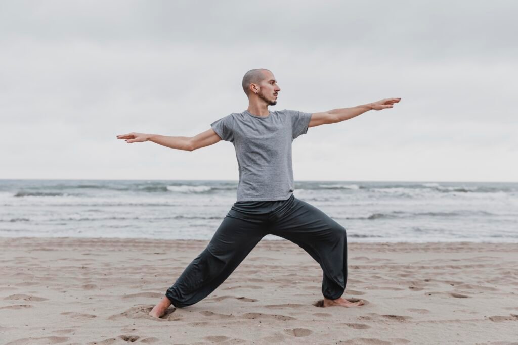 um jovem careca e de barba, praticando tai chi numa praia.