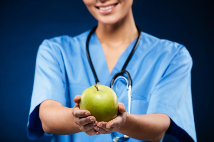 uma medica de jaleco azul oferecendo uma maçã com as duas mãos.
