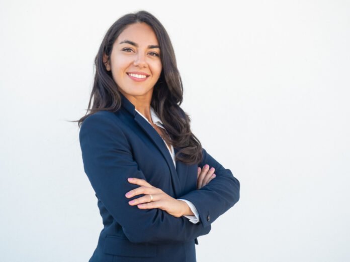 Uma bela mulher sorridente de cabelos longos vestida de um terninho executivo, em pose com os braços cruzados.