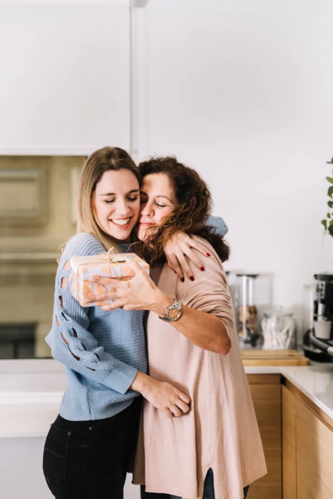 mae e filha abraçadas. a mãe segura uma caixa de presente quadrada.