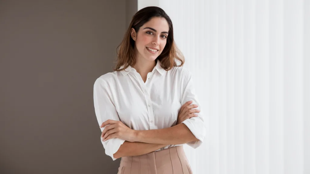 uma bela mulher sorridente, de camisa branca e saia marrom em pose com os braços cruzados.