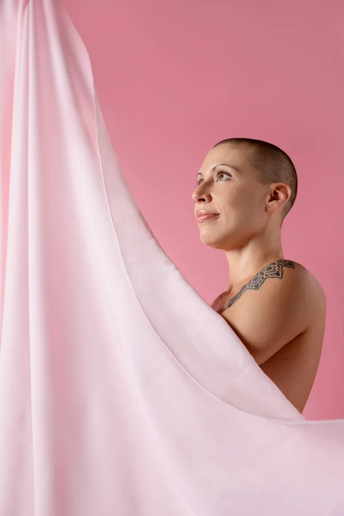 uma jovem de pela clara, careca, se cobrindo com um véu rosa claro num fundo rosa escuro.