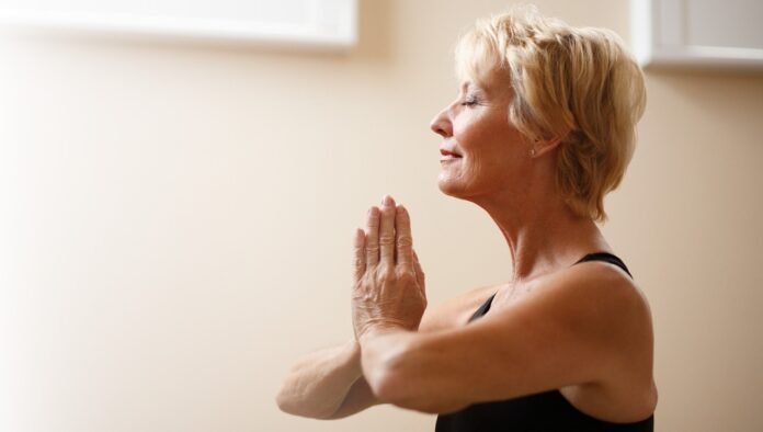 mulher loira, cabelos curtos, praticando yoga numa posição com as mãos postas.